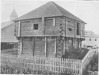 A weathered wooden building stands behind a white picket fence, with more modern structures visible in the background.