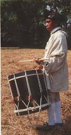 A man in traditional attire standing in a field, holding a drum.