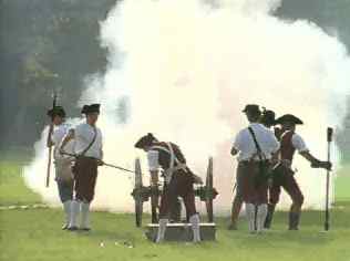 Historical reenactors in colonial-era clothing firing a cannon amidst a cloud of smoke on a grassy field.