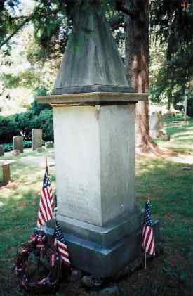 A tall, weathered obelisk monument stands in a peaceful cemetery, flanked by american flags, suggesting a site of remembrance for a notable figure or veteran.